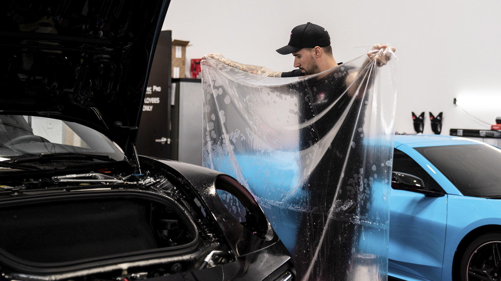 Ceramic Pro KAVACA PPF install in progress — installer prepping a slip-solution-soaked film panel beside a black Porsche and a blue McLaren